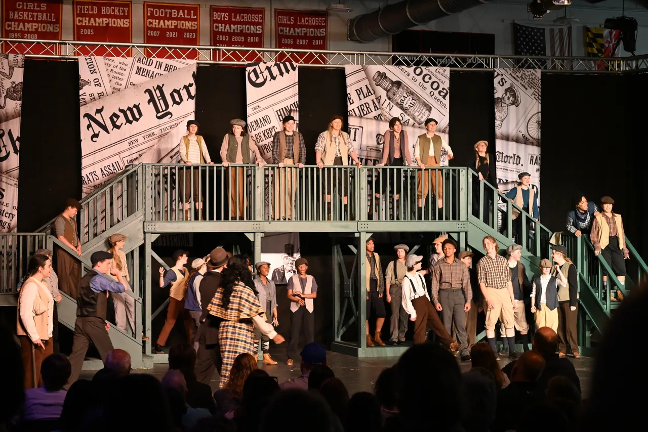 Students performing in the spring musical with a walkway and curtains with newspaper clippings.