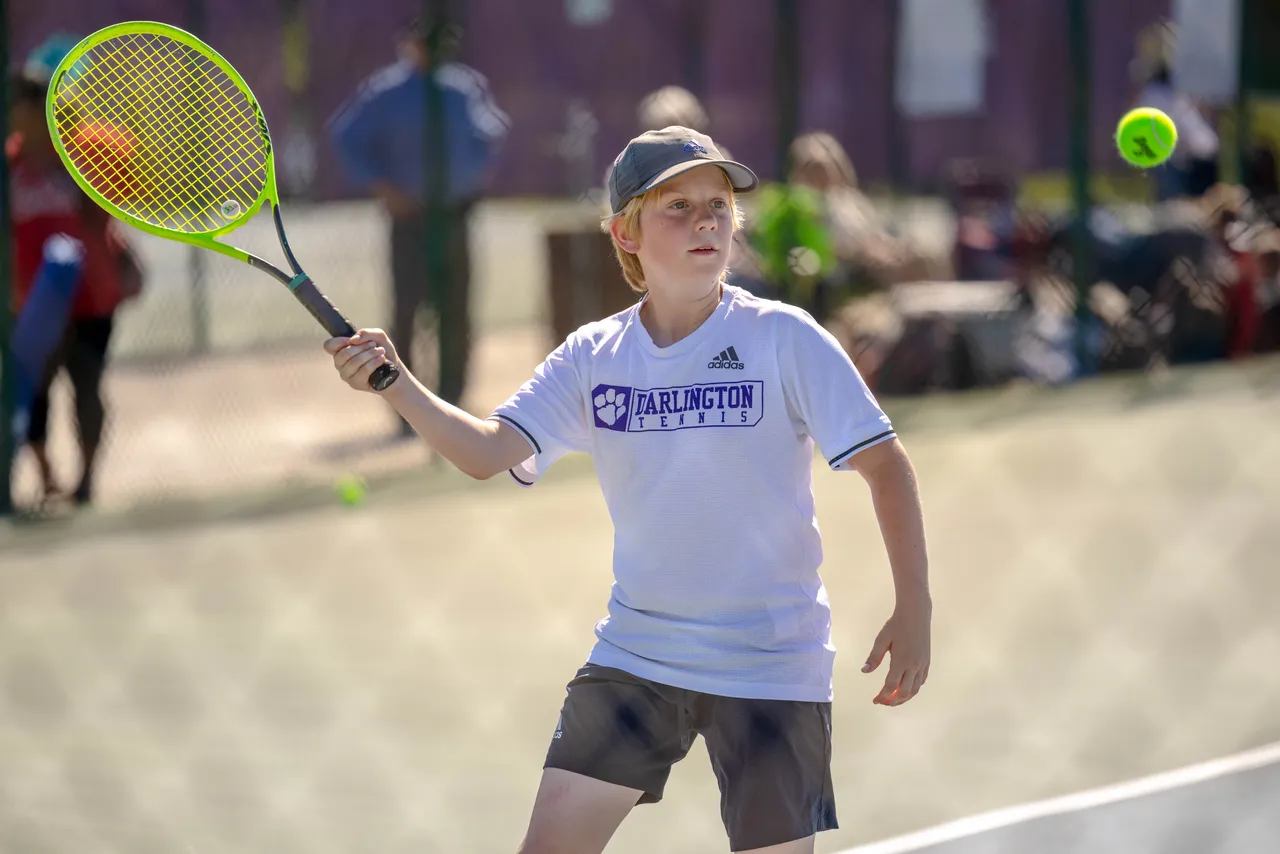 A male middle school tennis player swings his racket to return a tennis ball during a match.