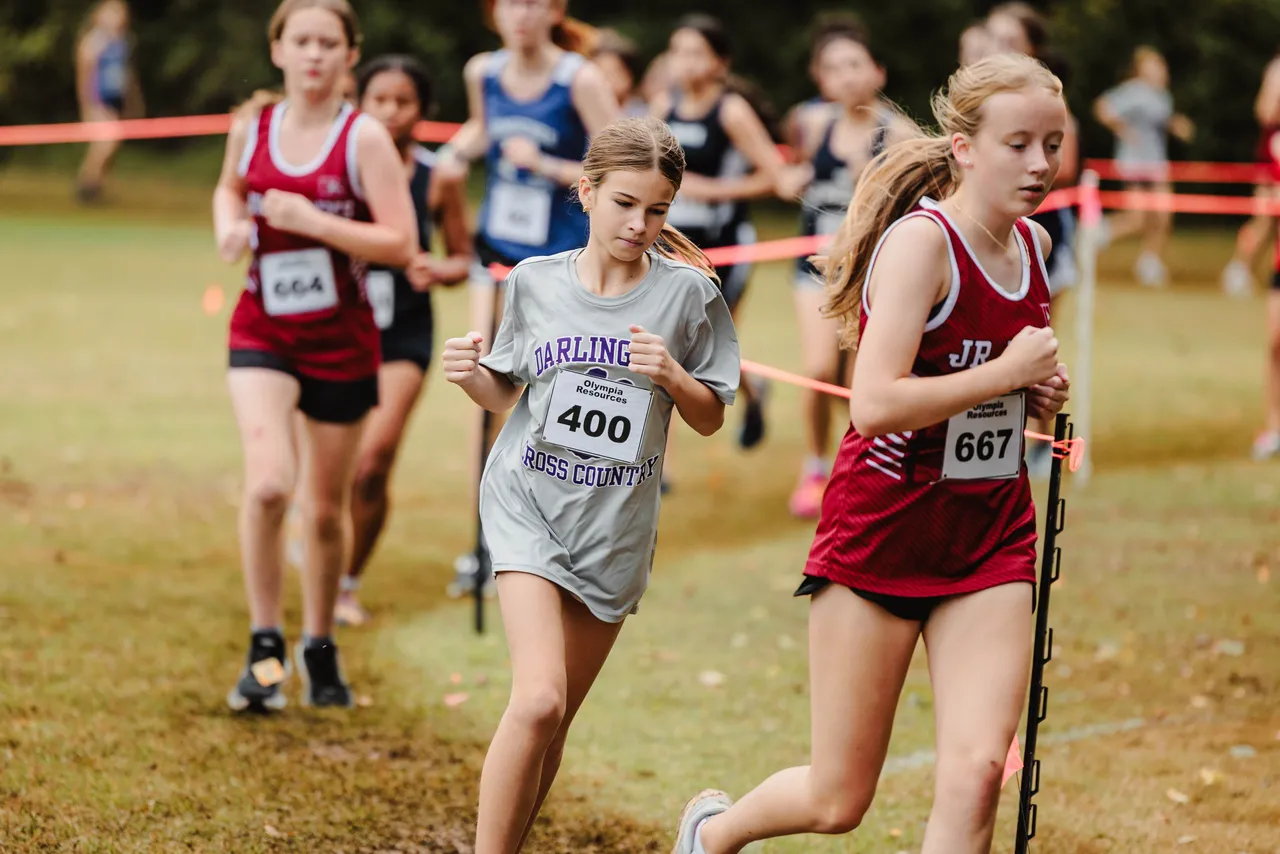 Female middle school JV cross country runners compete during a race on a cross country course.