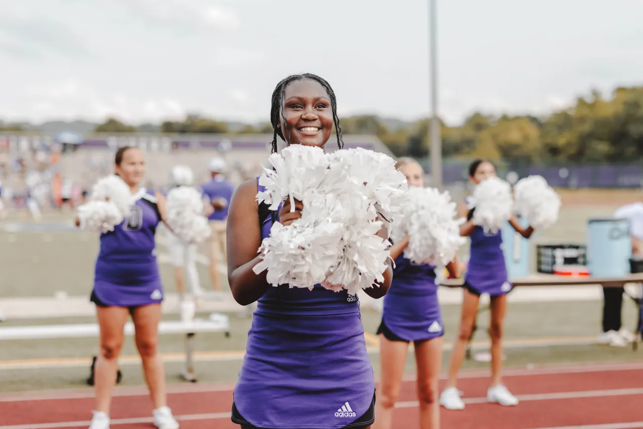 Female middle school football cheerleaders hold pom-poms and cheer on the sideline during a football game.