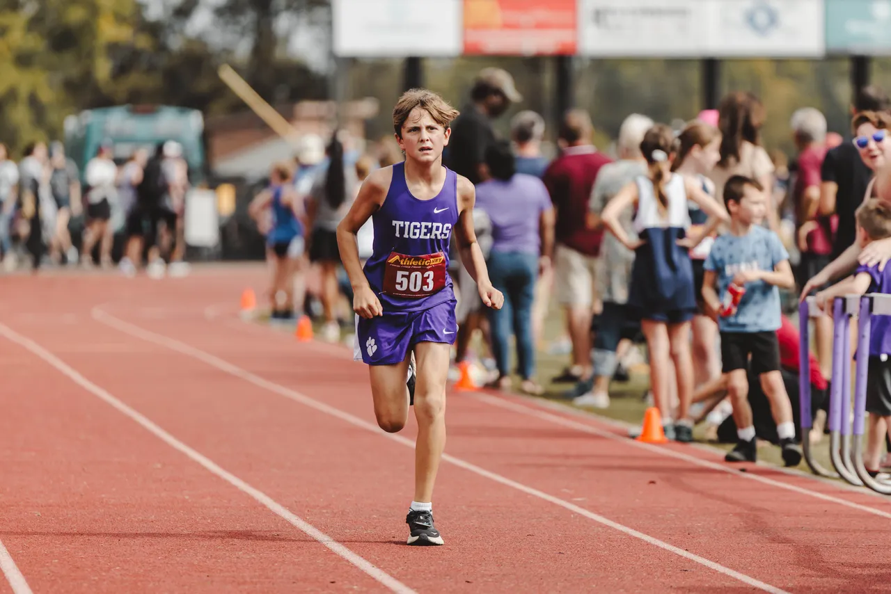 A male middle school JV cross country runner runs toward the finish line during a race on the track.
