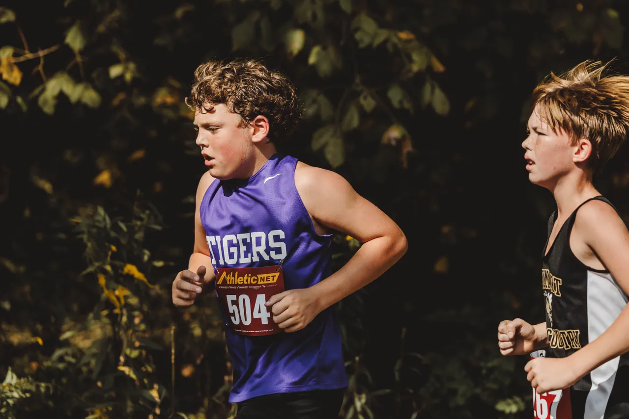 A male middle school cross country runner races alongside another runner during a cross country meet.