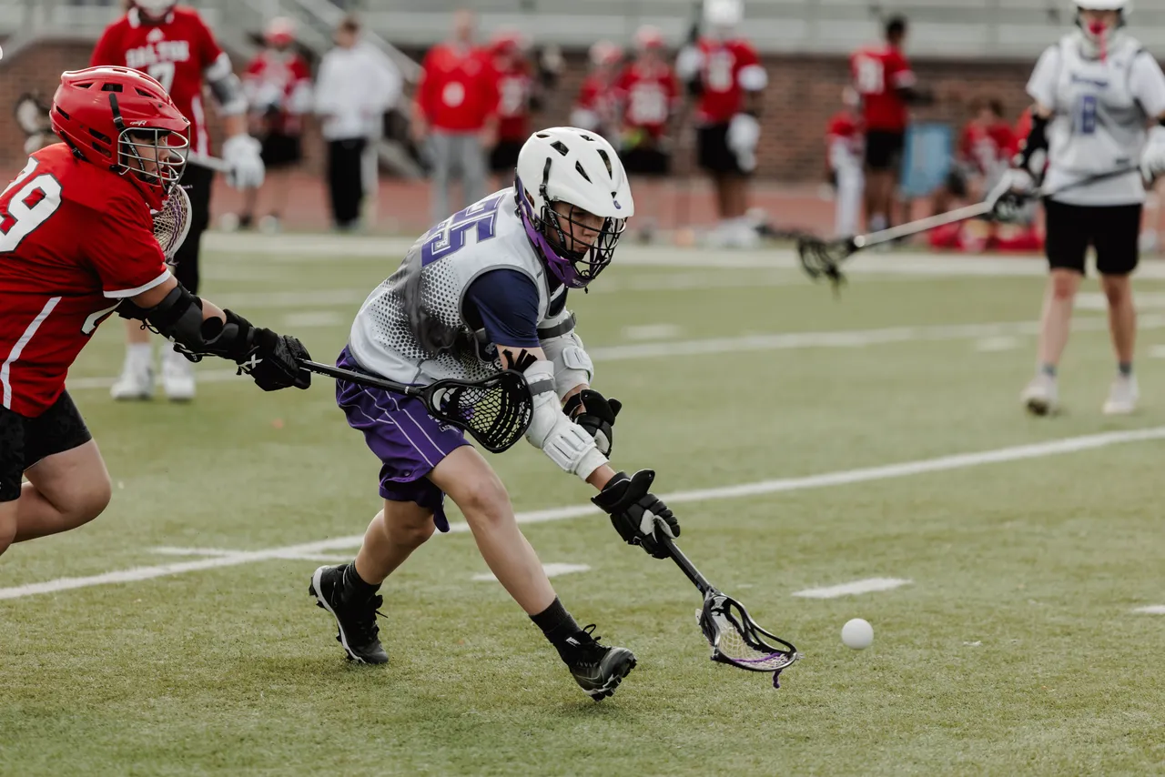 A male middle school lacrosse player carries the ball in his stick while an opposing player defends him during a game.