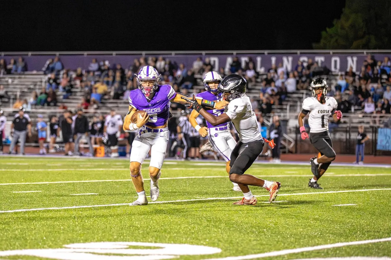 A Darlington football player in a purple jersey runs with the ball as he fends off a defender from the other team during a nighttime game, with teammates following the play and fans watching from the stands.