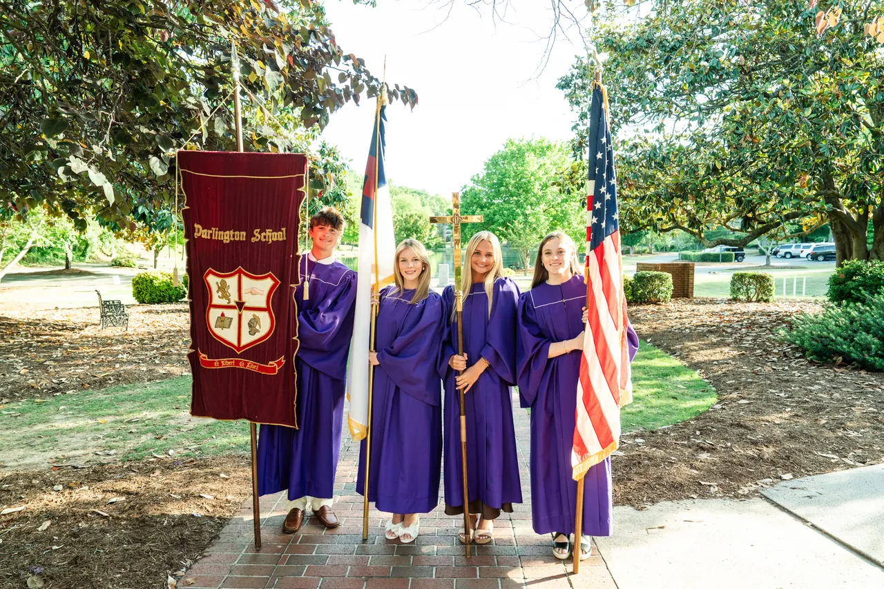 Four students in purple robes hold flags and a cross in front of Silver Lake at Darlington School.
