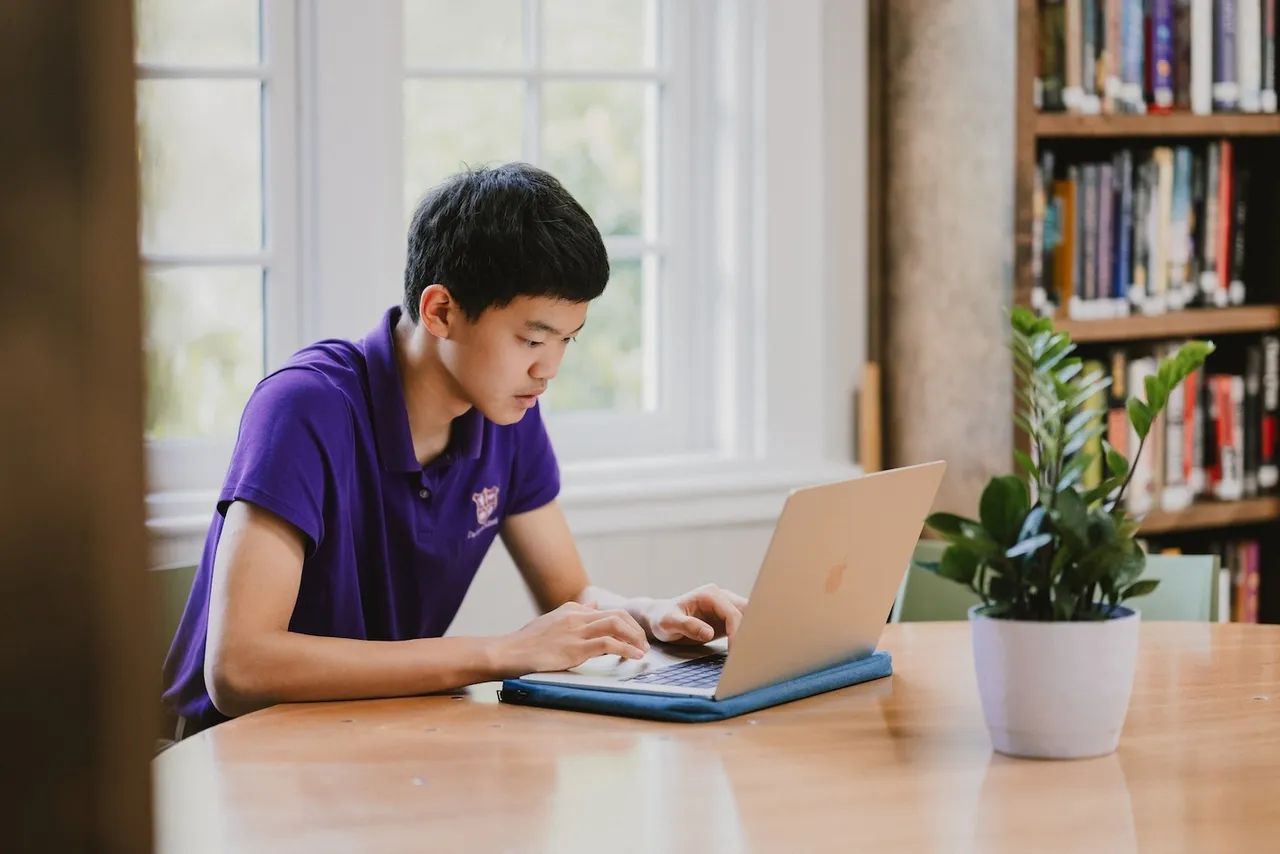 A male high school student works on his MacBook computer in the library between classes