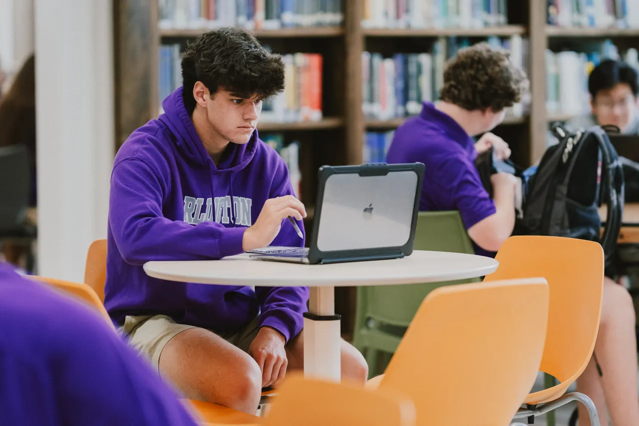 A male high school student works on his MacBook computer in the library between classes