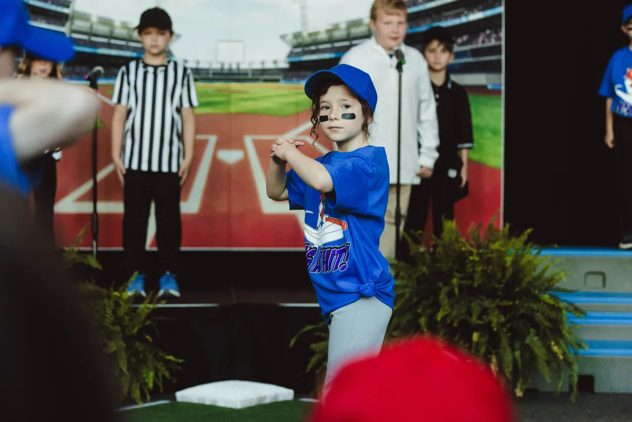 A female second grade student dressed in a baseball uniform performs in a class play themed around baseball