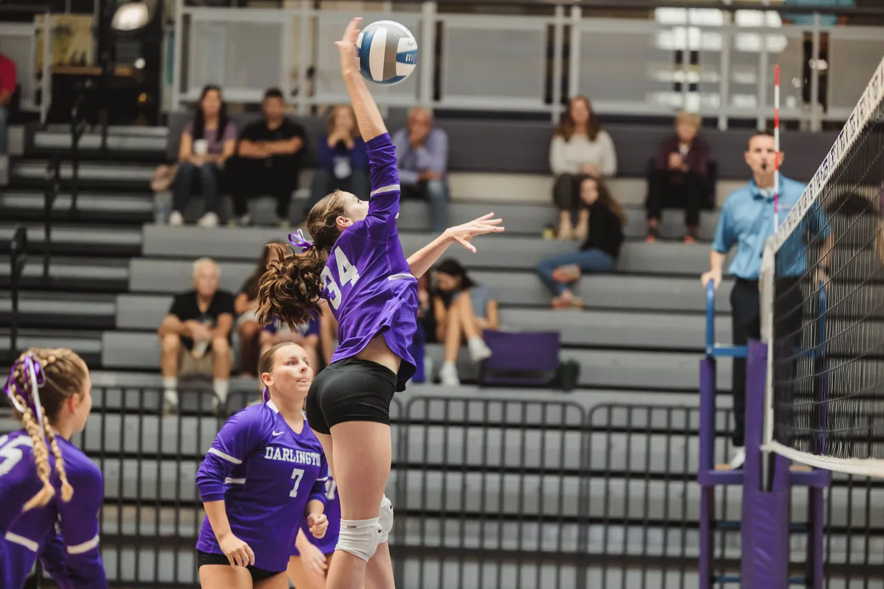A high school volleyball player jumps up to hit the ball at the net as her teammates look on