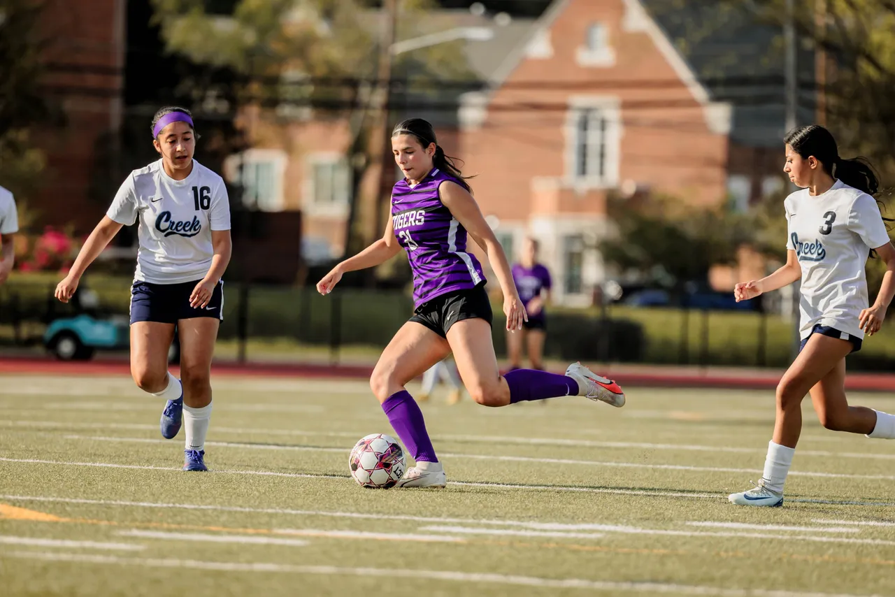 A female high school soccer player kicks the all as two opposing players run toward her