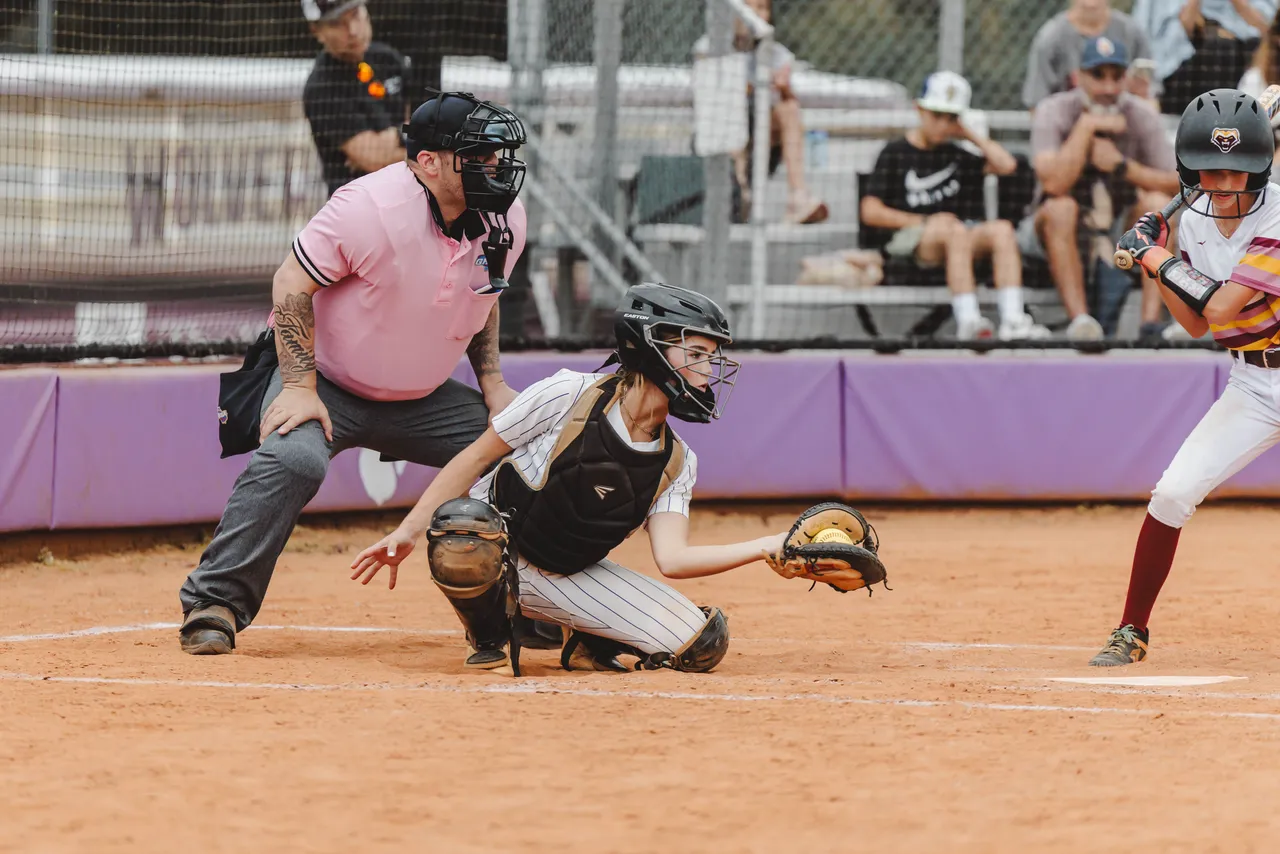 A high school softball catcher catches the ball in her glove as the umpire looks on behind her