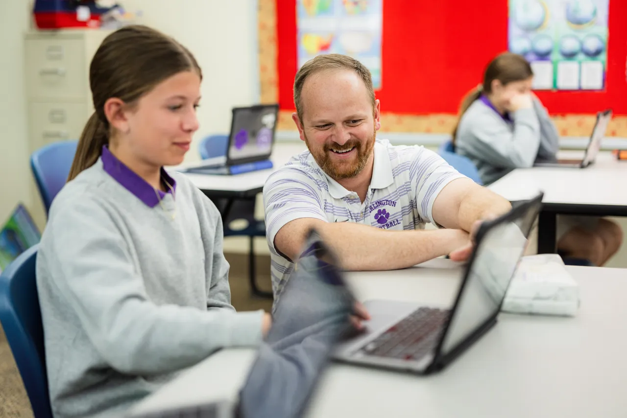 A male middle school teacher works with a female student on her MacBook computer in class