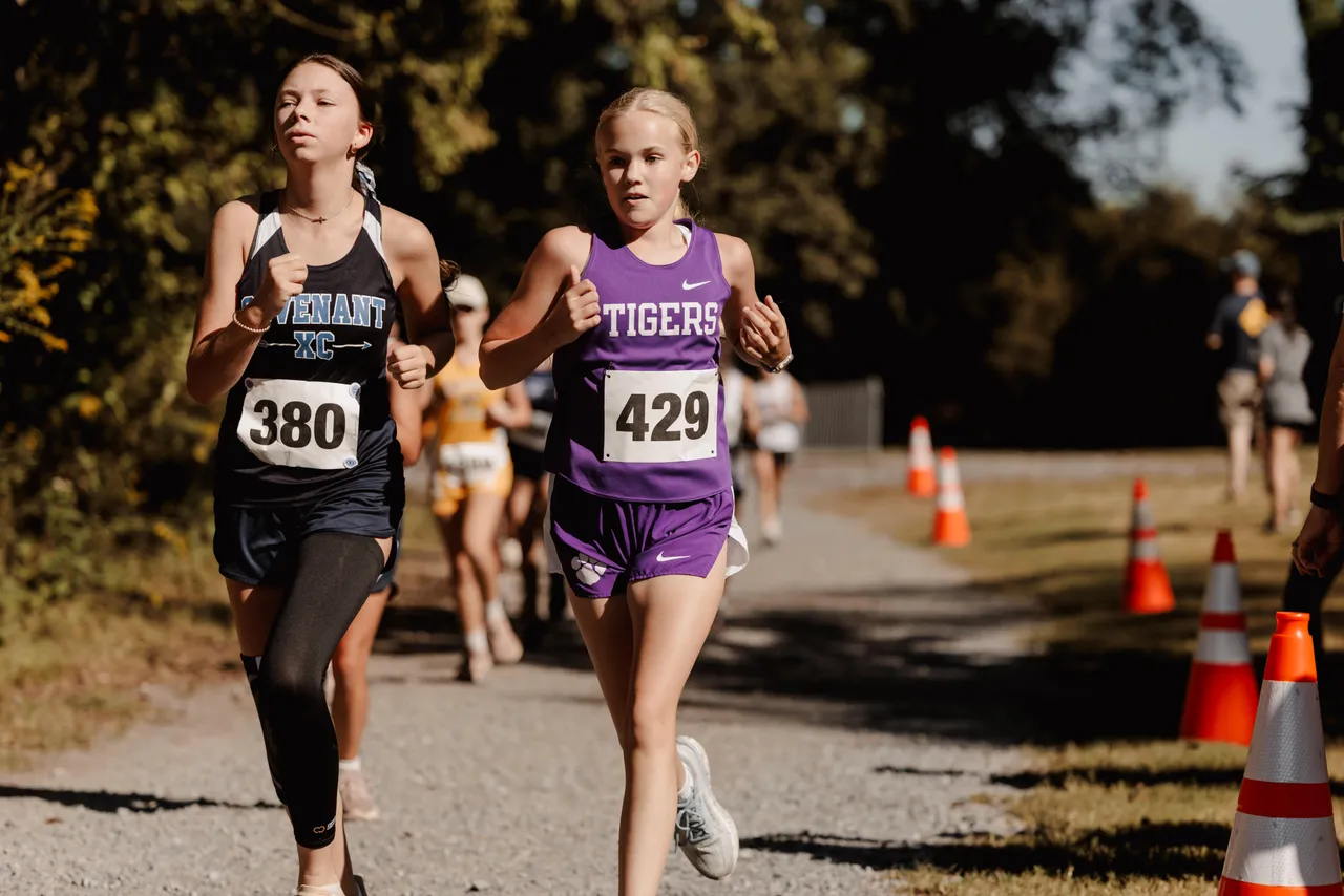 A female middle school cross country runner runs alongside another competitor during a race.