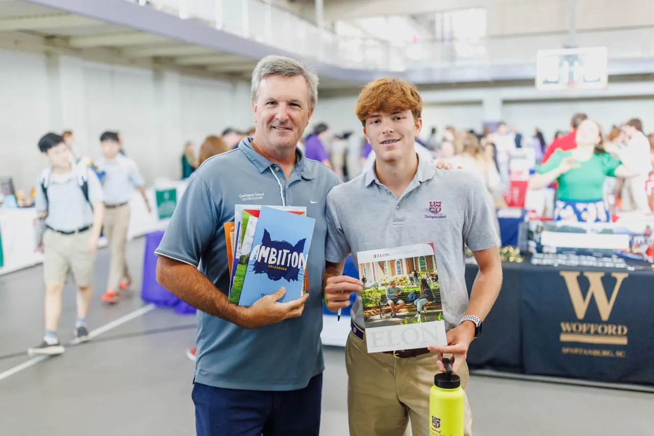 Male teacher holding college marketing materials stands next to a male high school student also holding college marketing materials during the college fair at Darlington School