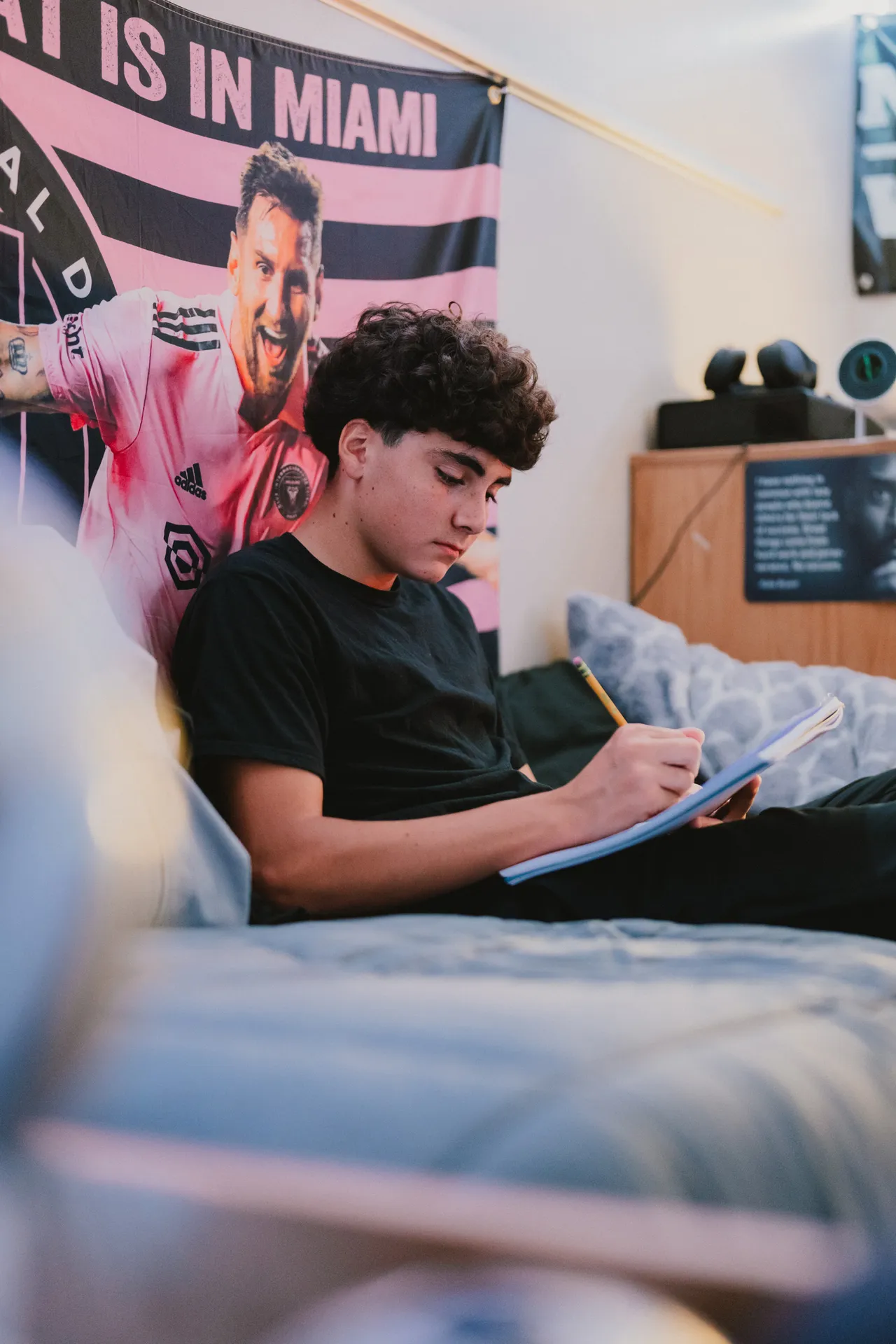 A male boarding student sits on his dorm room bed writing in a notebook