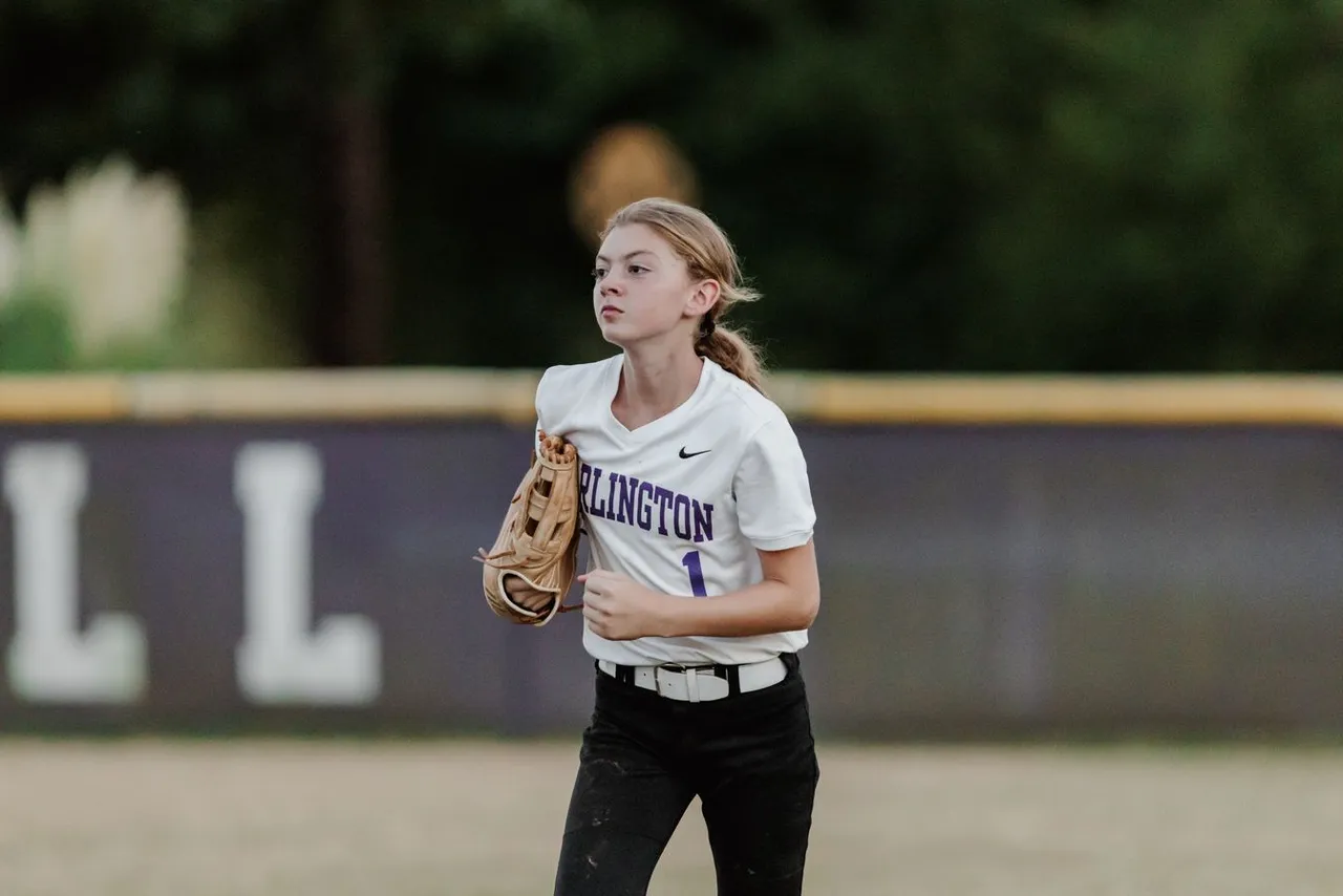 A female middle school softball player runs across the field with her glove during a game.