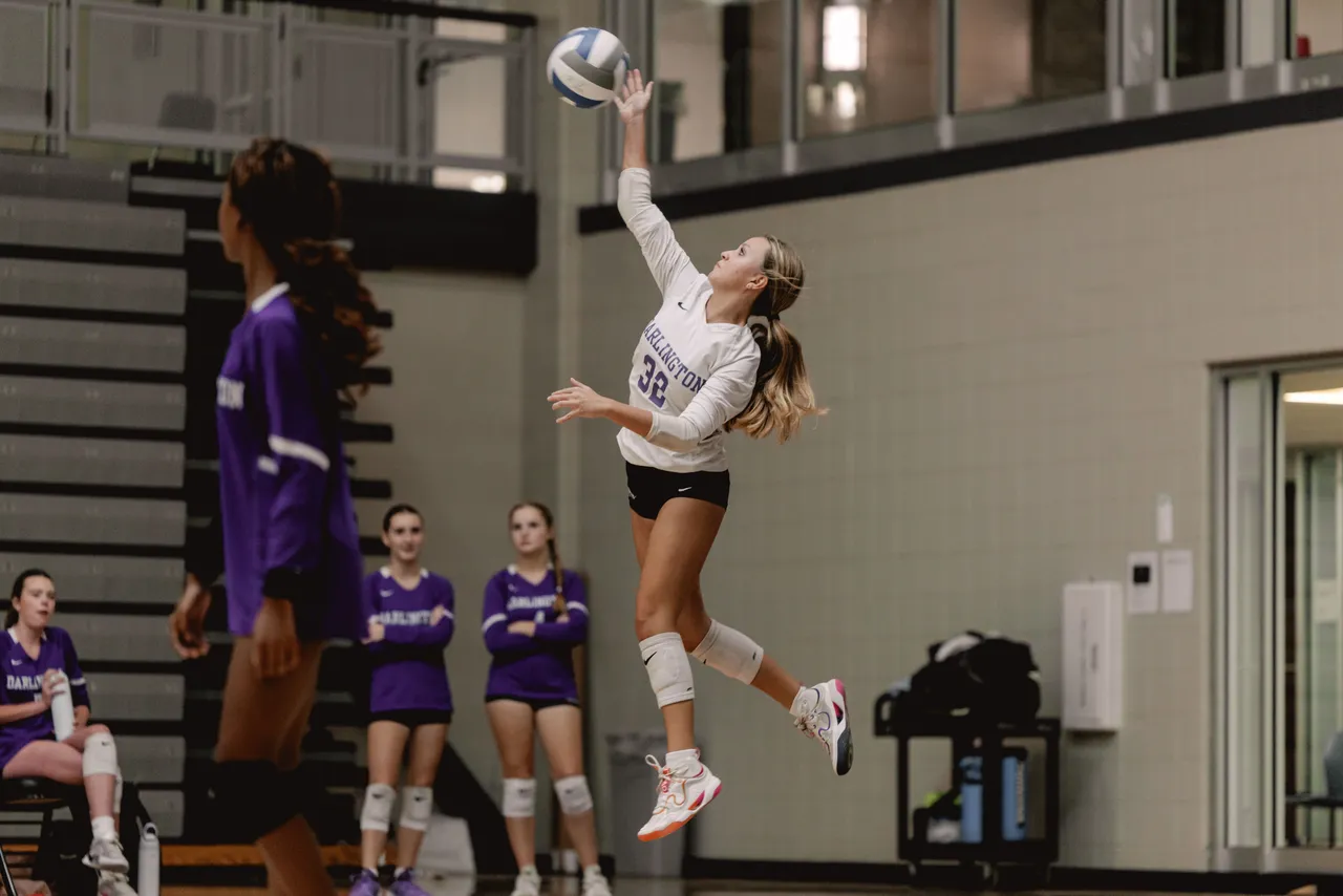 A female Darlington School JV volleyball player jumps to serve the ball during a match in the gym.