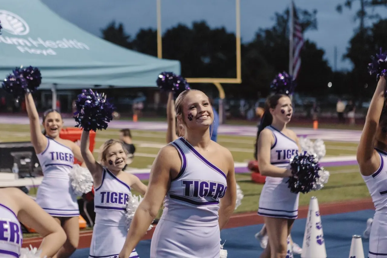 A high school cheerleader smiles while cheering on the sidelines during a football game.