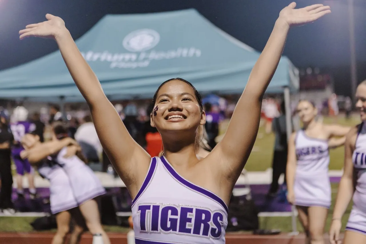 A high school cheerleader smiles with her hands in the air while cheering on the sidelines during a football game.