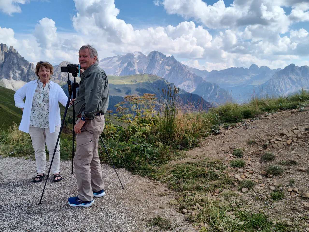 A woman and man smile at the camera while standing beside a film camera on a tripod in front of the Alpine mountain range