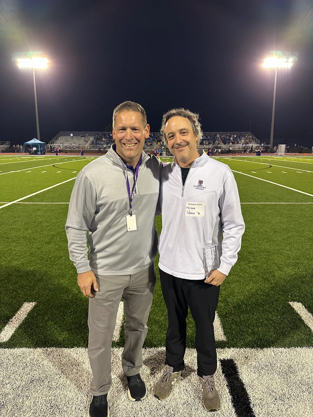 Two adult men stand side by side smiling on a football field