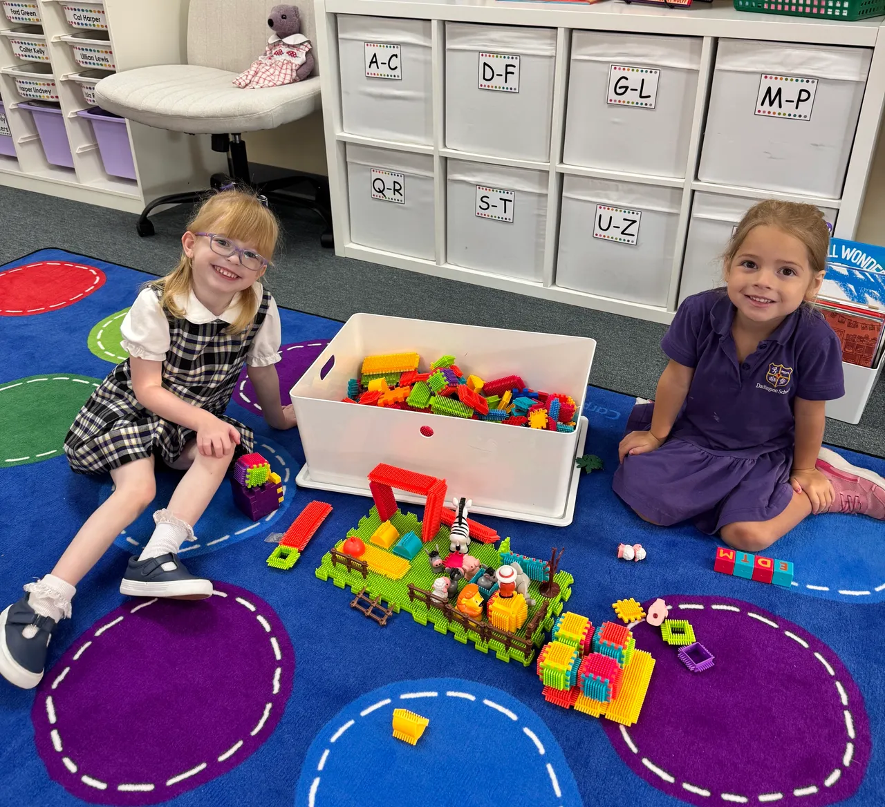 Two smiling female pre-K students sitting on a carpet with a farm scene that they have built using blocks and farm animal toys