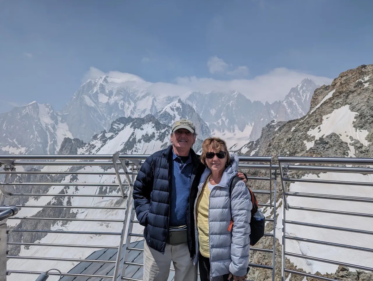 A man wearing a ballcap and a woman wearing sunglasses, both in puffer coats, stand in front of the snowy Alpine mountains in France