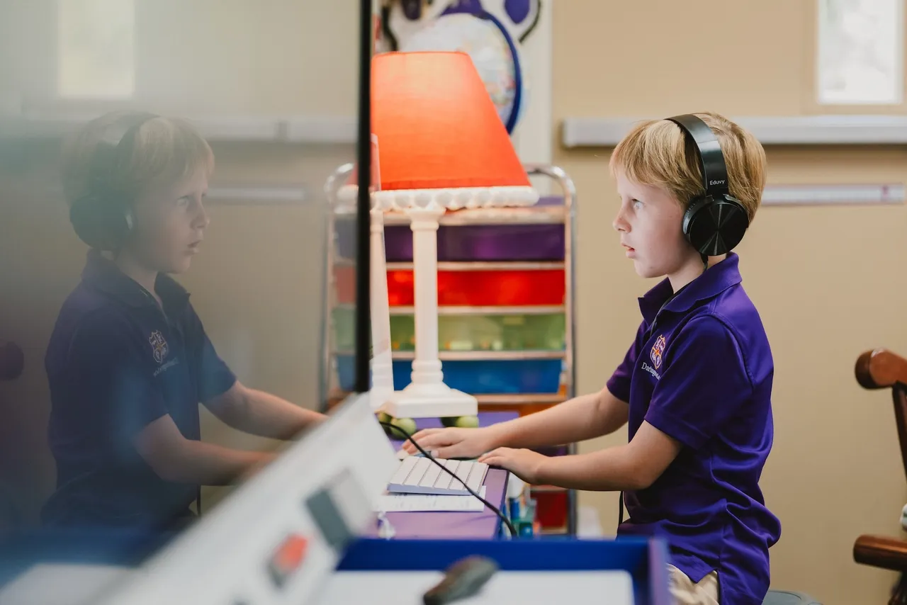 Male primary grades student works on a computer in his classroom while wearing headphones