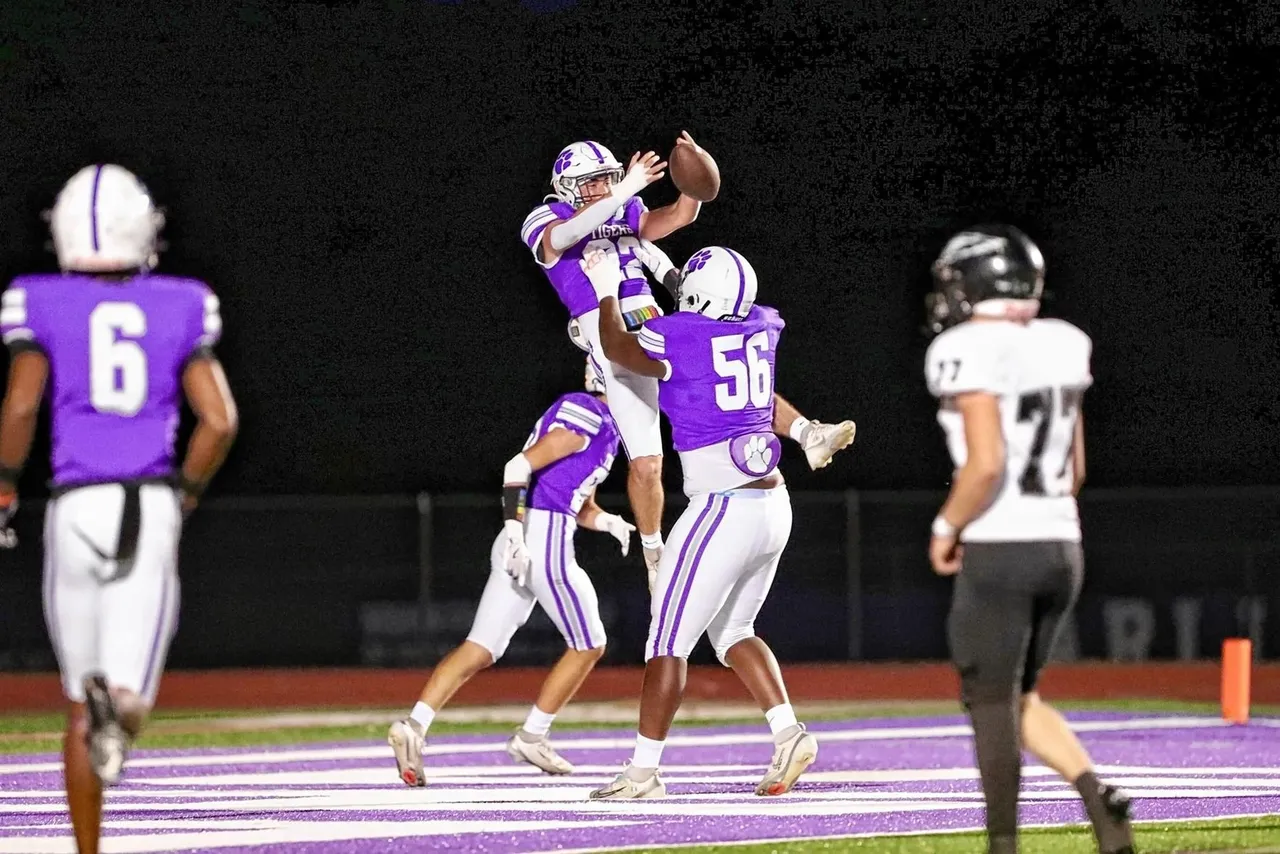 A Darlington football player in a purple jersey leaps into the air to celebrate a touchdown as teammate No. 56 lifts him up in the end zone during a nighttime game.