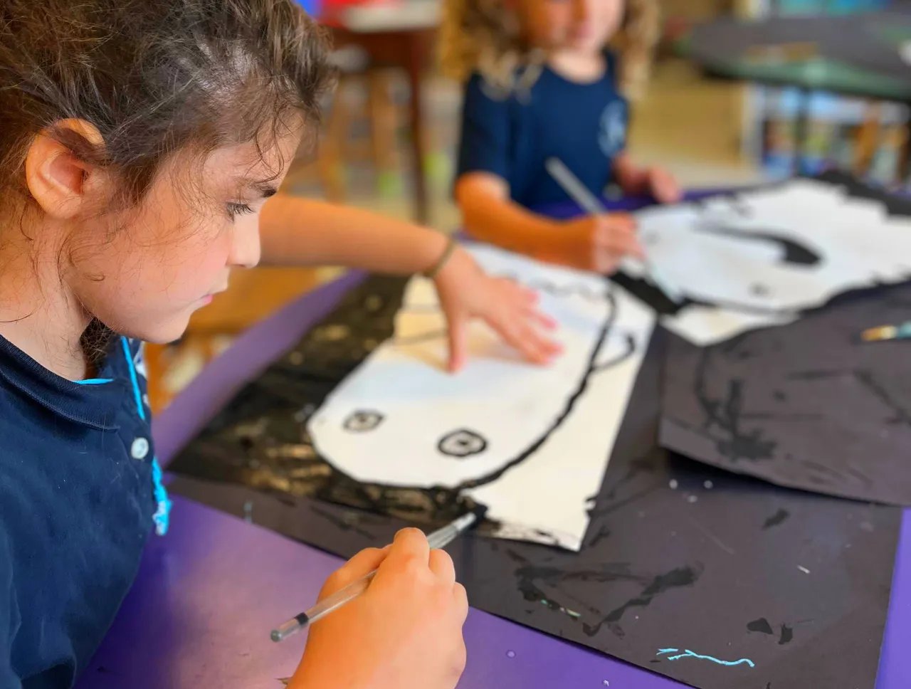 Two Mason Prep students work on a classroom art project, tracing and coloring large paper shapes at a table.
